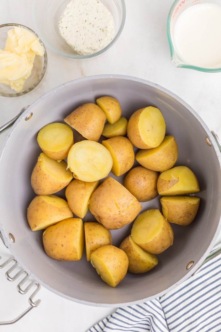 A pot filled with boiled, halved potatoes sits on a white surface. Surrounding the pot are small bowls holding butter, a block of cream cheese, and a measuring cup of milk. A striped cloth is partially visible.
