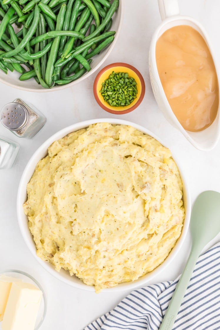 A bowl of creamy mashed potatoes sits on a white surface, surrounded by a dish of green beans, a small bowl of chopped chives, a gravy boat, a butter dish, salt and pepper shakers, and a striped napkin.