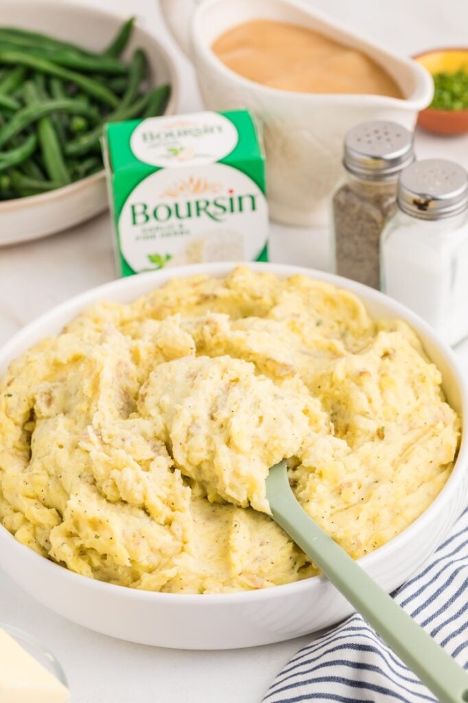 A bowl of creamy mashed potatoes with herbs is being served with a green spatula. In the background, there are green beans, a box of Boursin cheese, gravy, salt, pepper, and a striped napkin.