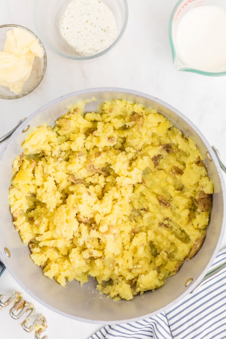 A pot filled with roughly mashed yellow potatoes sits on a white surface, surrounded by bowls of butter, a creamy mixture, and a cup of milk. A striped cloth napkin is partially visible.