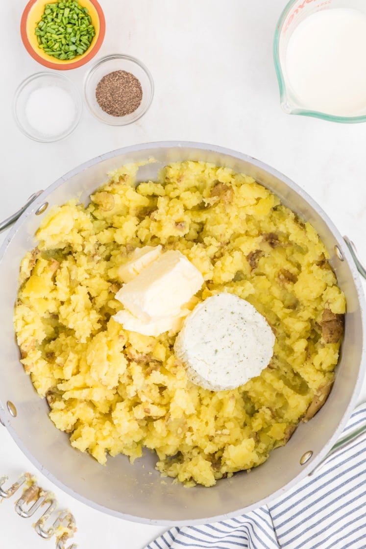 A pot of partially mashed potatoes with butter and a round of herb cheese on top, surrounded by bowls of chives, salt, pepper, and a measuring cup of milk. A striped towel is nearby.
