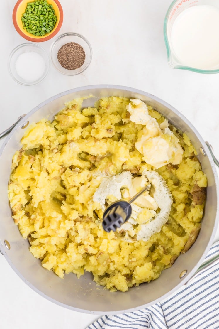 A pot of partially mashed potatoes with butter, cream, and a masher. Surrounding the pot are small bowls of chives, salt, pepper, and a measuring cup of milk. A striped towel sits nearby on the countertop.
