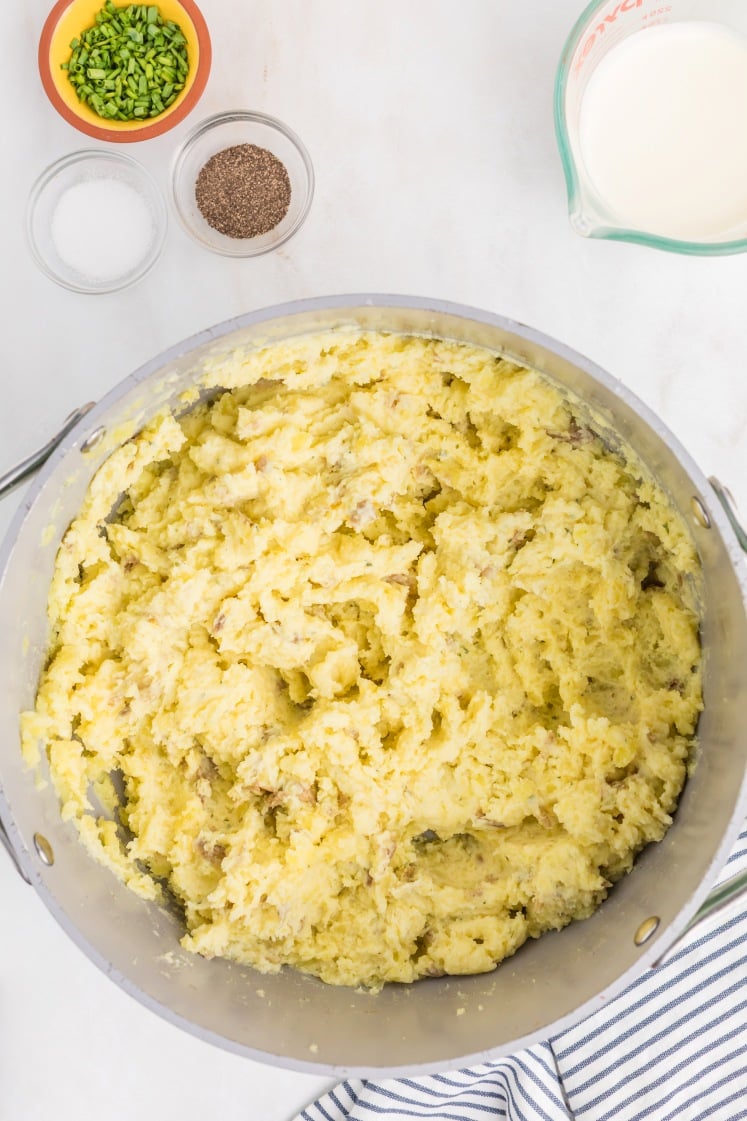 A large pot filled with mashed potatoes sits on a white surface. Nearby are small bowls of chopped chives, salt, pepper, and a measuring cup of milk. A striped towel is partially visible in the corner.