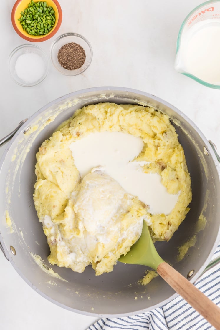 A pot of mashed potatoes with milk being stirred in by a green spatula. Nearby are small bowls of chopped chives, salt, pepper, and a measuring cup of milk on a white surface.