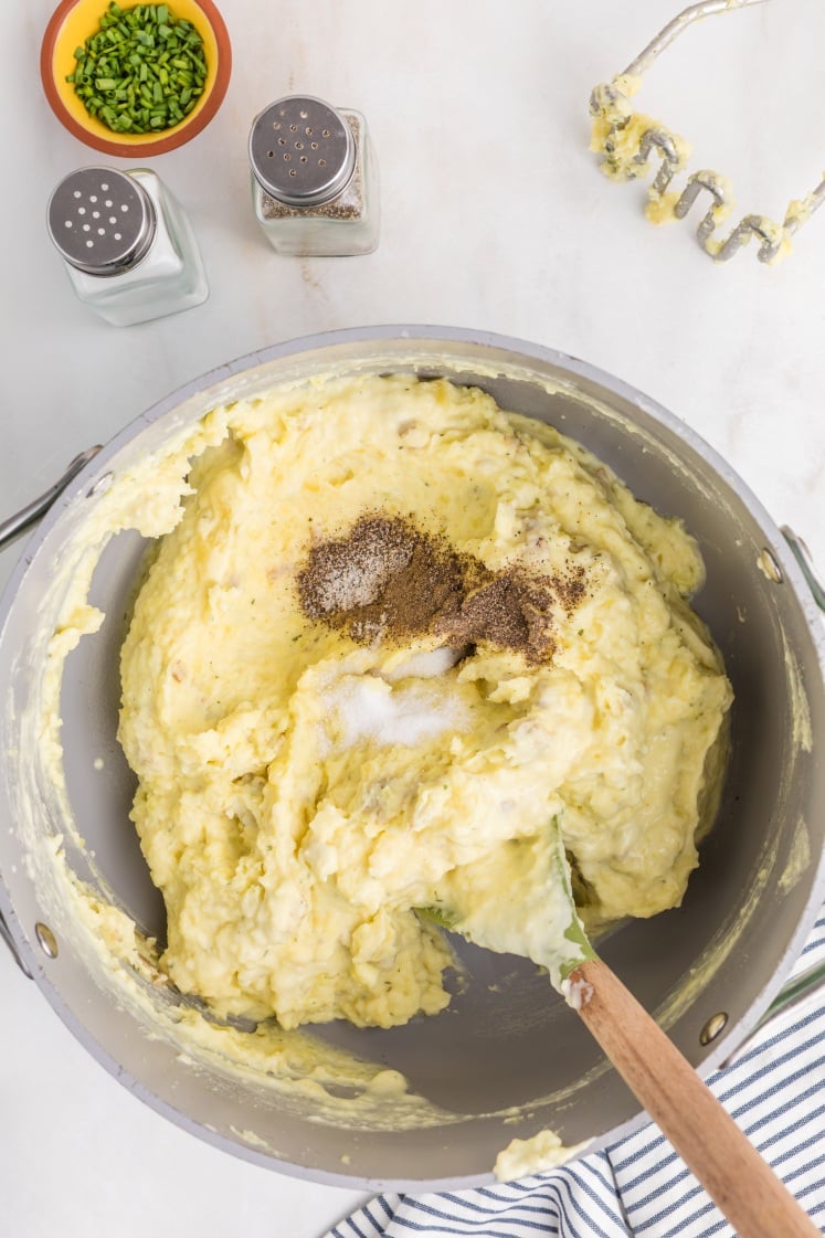 A mixing bowl filled with mashed potatoes being seasoned with salt and pepper, with a wooden spoon for stirring. Nearby are a hand masher, salt and pepper shakers, chopped chives, and a striped kitchen towel.