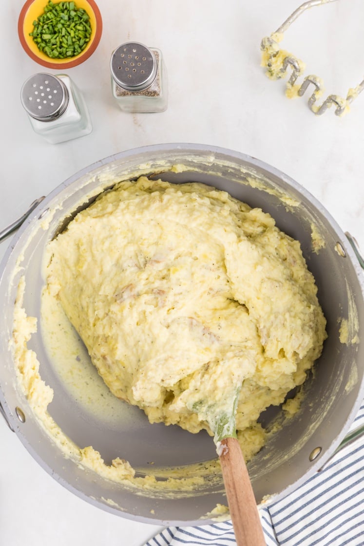 A large pot of creamy mashed potatoes with a wooden spoon, next to salt and pepper shakers, a bowl of chopped chives, a potato masher, and a striped kitchen towel on a white surface.