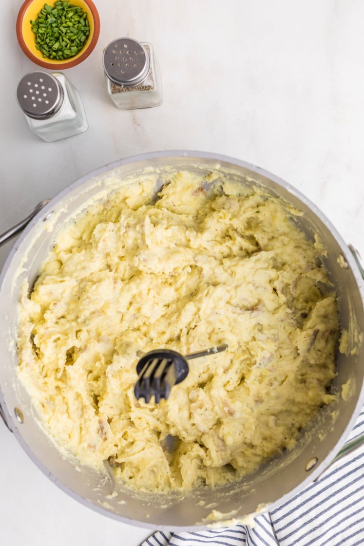 A large pot filled with mashed potatoes and a hand masher sits on a white surface. Nearby are a bowl of chopped chives and salt and pepper shakers. A striped cloth is partially visible at the bottom.