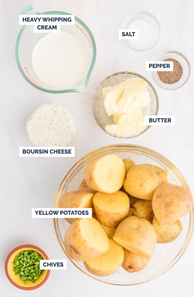 Labeled ingredients for mashed potatoes: a bowl of yellow potatoes, a cup of heavy whipping cream, small bowls of salt, pepper, and butter, Boursin cheese, and chopped chives on a light background.