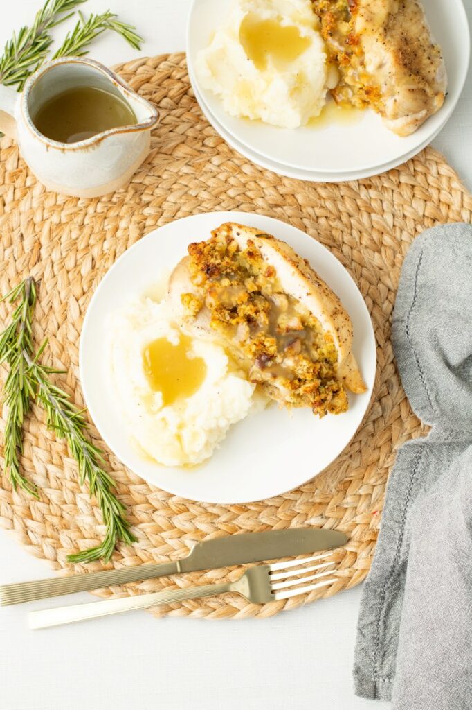 A plate with mashed potatoes topped with gravy and a serving of stuffing-covered chicken breast, placed on a woven mat with rosemary sprigs, gold utensils, and a small pitcher of gravy nearby.