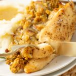 A close-up of a plate with stuffed chicken breast being sliced, served with mashed potatoes and brown gravy. The stuffing appears to include bread crumbs and herbs. The food is on a white plate atop a woven placemat.