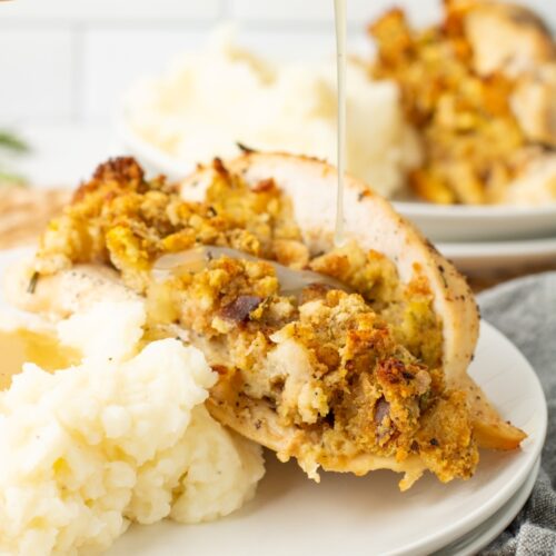 A close-up of a plate with mashed potatoes and stuffed chicken breast, topped with bread stuffing. Gravy is being poured over the chicken. Another plate with the same meal is blurred in the background.