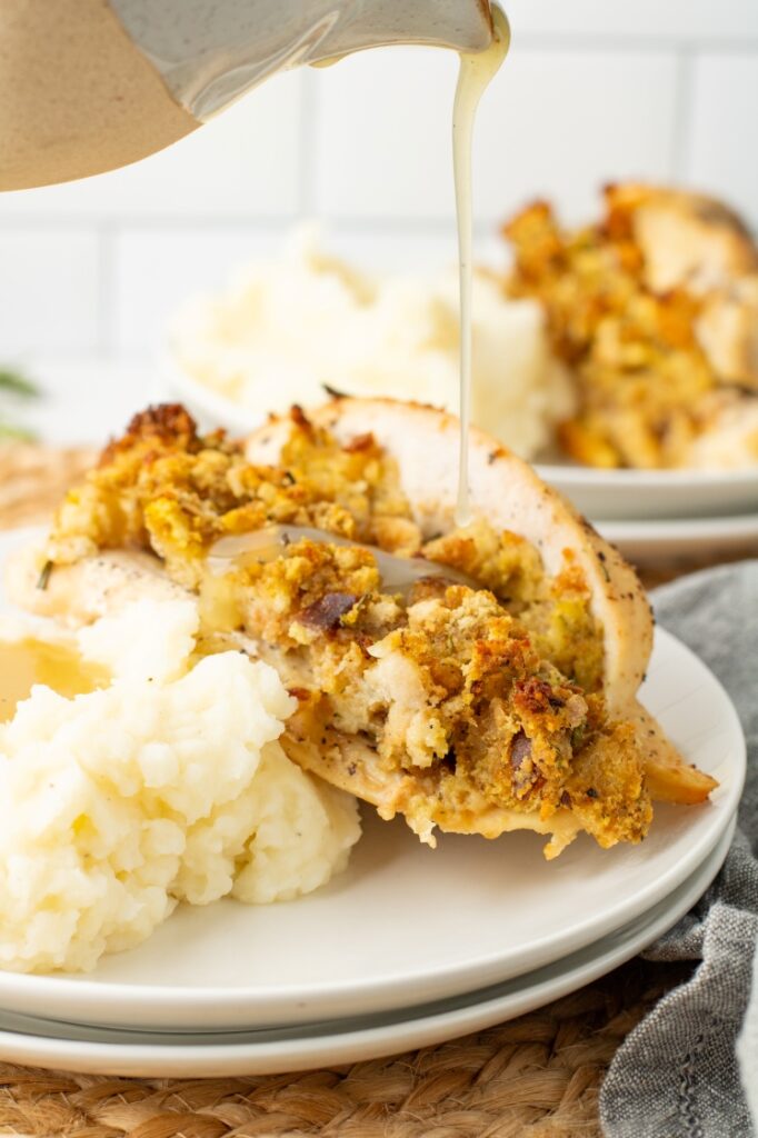 A close-up of a plate with mashed potatoes and stuffed chicken breast, topped with bread stuffing. Gravy is being poured over the chicken. Another plate with the same meal is blurred in the background.