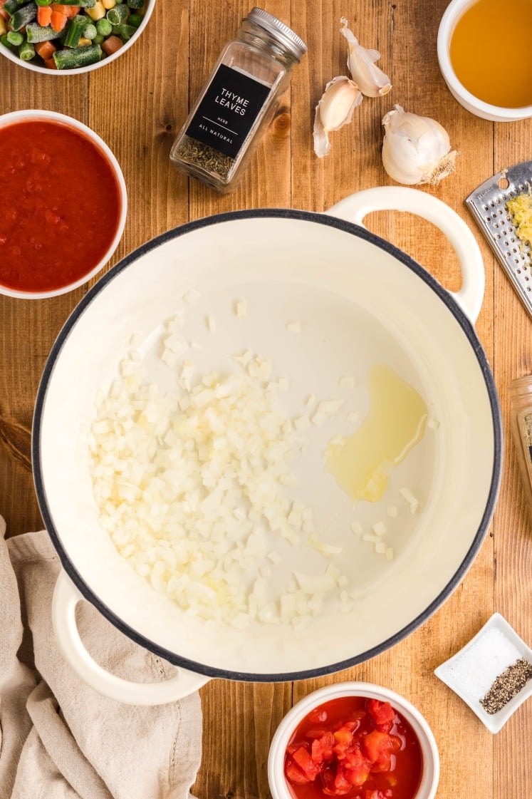 Top-down view of a soup pot with diced onions and oil, surrounded by ingredients including canned tomatoes, tomato sauce, garlic, grated garlic, thyme leaves, broth, salt, and pepper on a wooden surface.