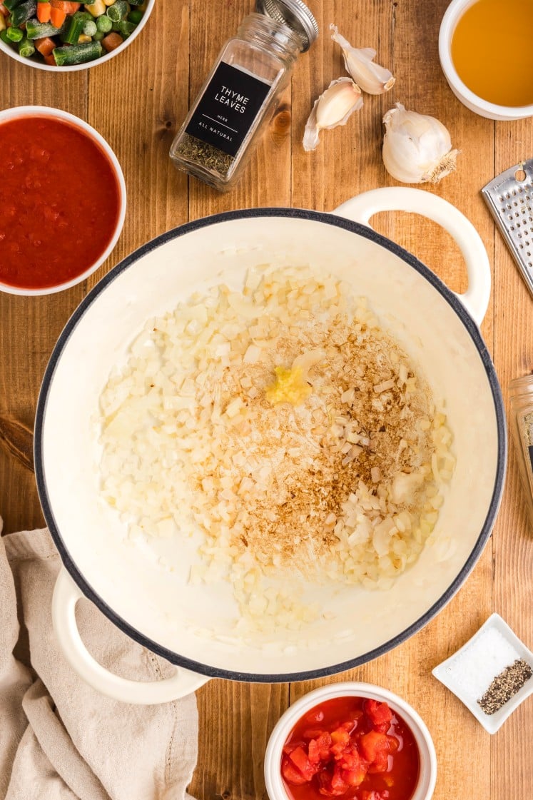 Top-down view of a pot filled with sautéing diced onions, spices, and minced garlic, surrounded by ingredients including thyme, garlic cloves, broth, tomato sauce, diced tomatoes, and seasonings on a wooden surface.