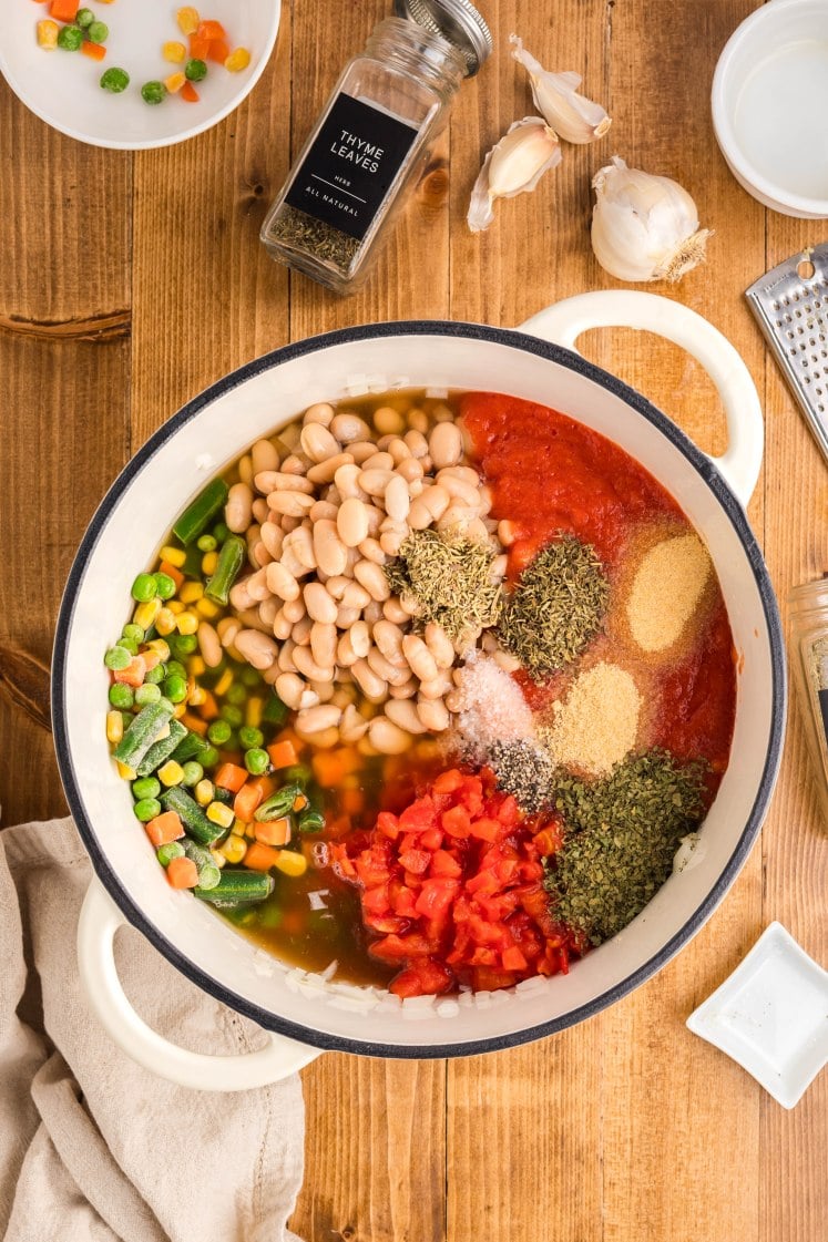 view of a soup pot filled with uncooked ingredients, including white beans, frozen mixed vegetables, diced tomatoes, tomato sauce, broth, and various dried herbs and seasonings, surrounded by thyme, garlic, and bowls on a wooden surface.
