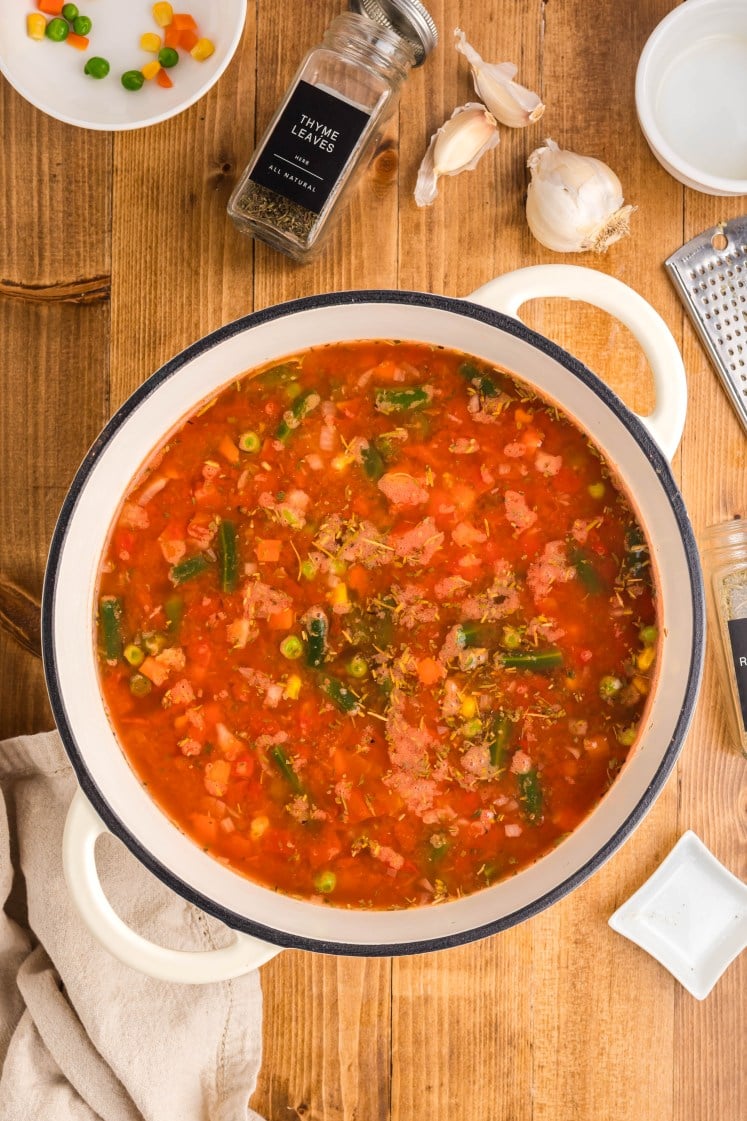 A pot of simmering vegetable soup with tomatoes, mixed vegetables, herbs, and broth, surrounded by thyme, garlic, and small bowls on a wooden surface.