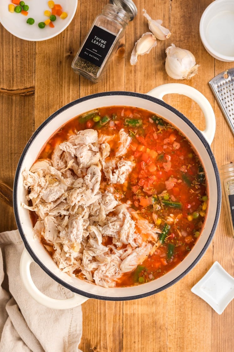 Top-down view of a pot of vegetable soup with shredded cooked chicken added on top, surrounded by thyme, garlic, bowls, and mixed vegetables on a wooden surface.