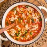 Top-down view of a pot of hearty chicken vegetable soup with shredded chicken, peas, corn, carrots, green beans, and tomatoes, stirred with a wooden spoon and surrounded by crackers, herbs, and garlic on a wooden surface.