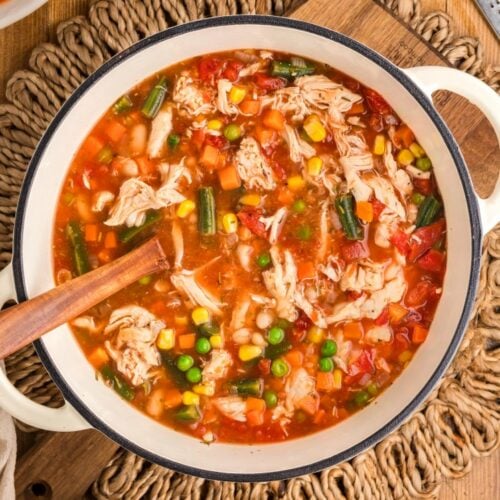 Top-down view of a pot of hearty chicken vegetable soup with shredded chicken, peas, corn, carrots, green beans, and tomatoes, stirred with a wooden spoon and surrounded by crackers, herbs, and garlic on a wooden surface.