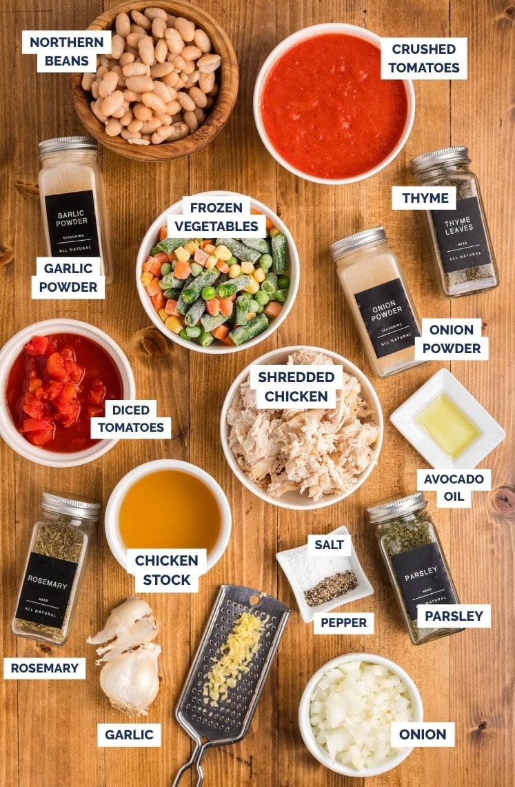 Top-down view of chicken vegetable soup ingredients arranged on a wooden surface, including northern beans, crushed tomatoes, diced tomatoes, frozen mixed vegetables, shredded chicken, chicken stock, avocado oil, chopped onion, minced garlic, garlic powder, onion powder, thyme, rosemary, parsley, salt, and pepper, each labeled with text.