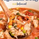 Close-up of a ladle lifting a hearty scoop of chicken vegetable soup from a pot, filled with shredded chicken, tomatoes, beans, corn, peas, and carrots, with the text “Chicken Vegetable Soup - Real Balanced" displayed above.