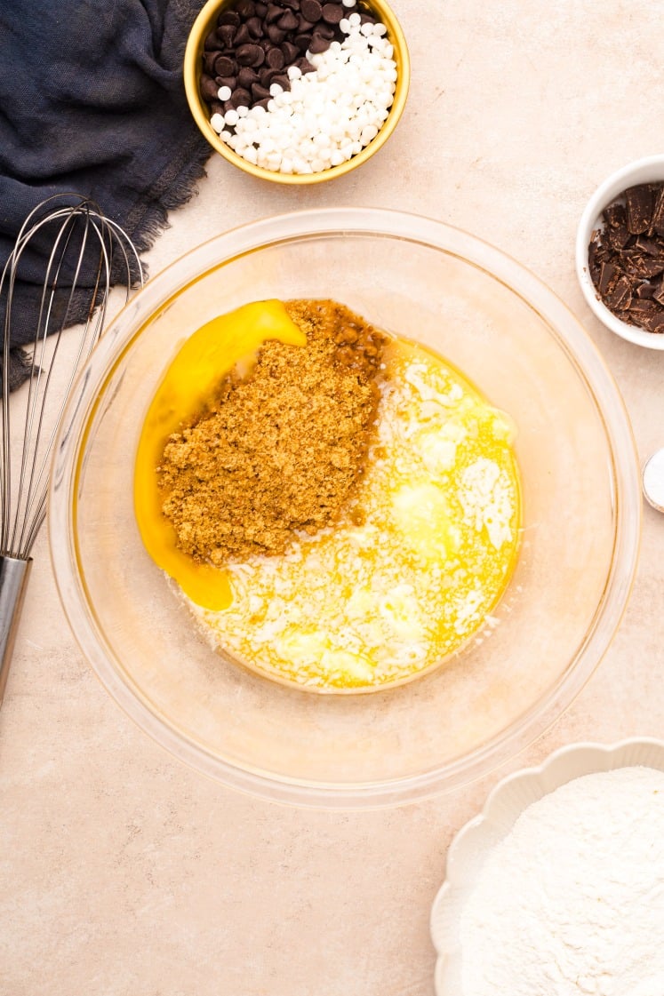 Overhead view of a glass mixing bowl containing melted butter, brown sugar, and egg yolks on a beige countertop. Nearby are bowls of chocolate chips, white chocolate chips, flour, and a whisk.