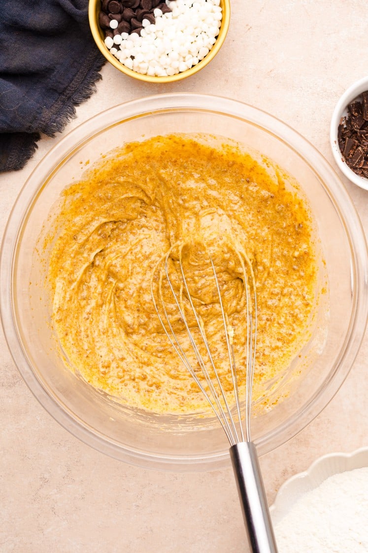 Glass mixing bowl with a whisk stirring a thick golden-brown mixture of eggs, butter, and brown sugar on a beige countertop, surrounded by bowls of chocolate chips and flour, part of the cookie dough preparation process.