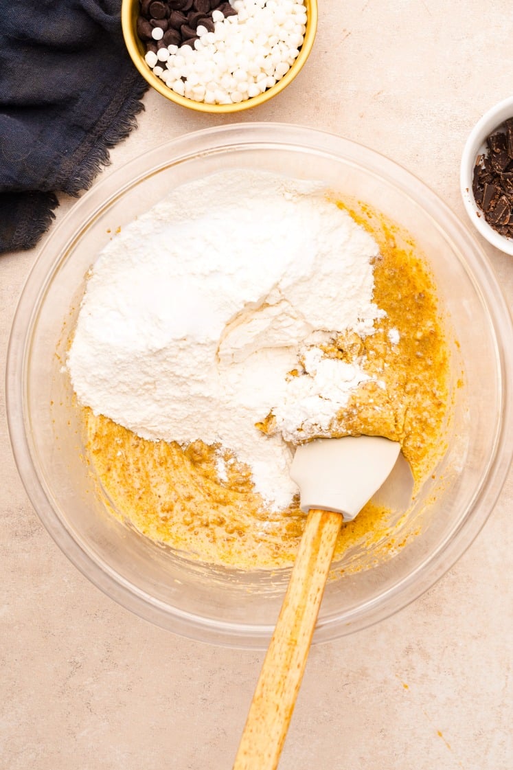 Glass mixing bowl with flour added to a creamed butter and sugar mixture, ready to be combined using a white spatula with a wooden handle.