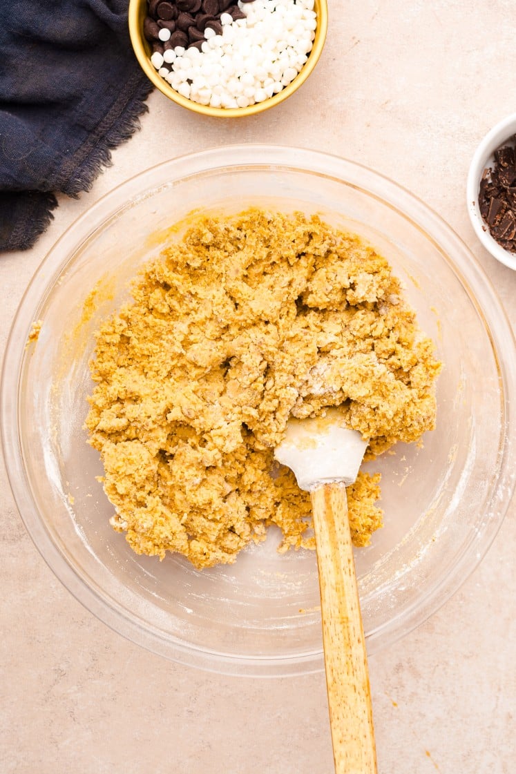Glass mixing bowl filled with thick, golden cookie dough being mixed with a white spatula with a wooden handle on a beige countertop, showing the dough just combined and ready for add-ins.