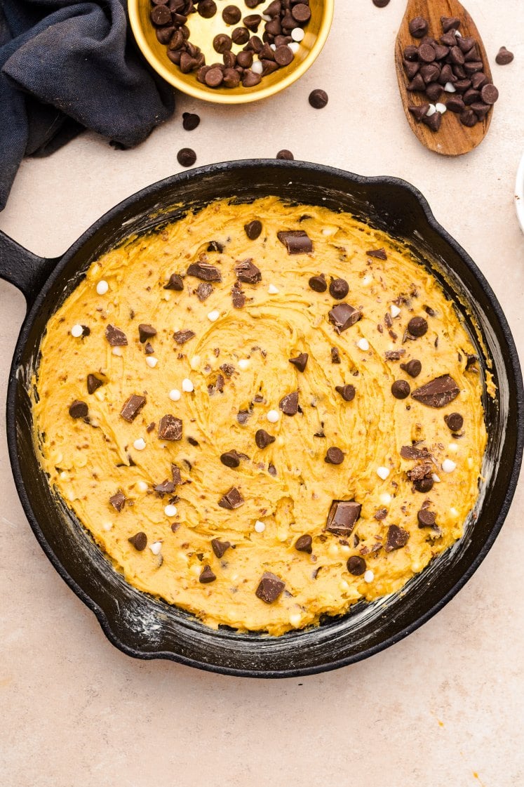 A cast iron skillet filled with unbaked cookie dough topped with chocolate chips and chocolate chunks, resting on a light-colored surface.