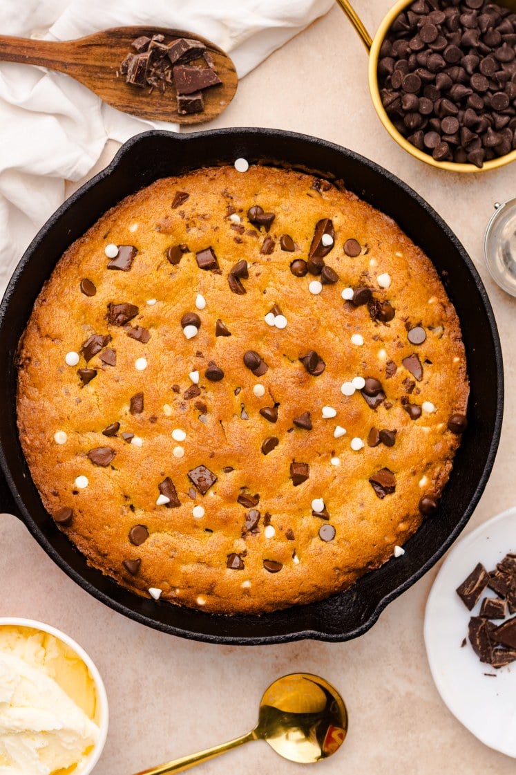 A large chocolate chip cookie baked in a cast iron skillet, topped with chocolate chunks and white chips. Surrounding the skillet are bowls of chocolate chips, a scoop of ice cream, and a wooden spoon with chocolate pieces.