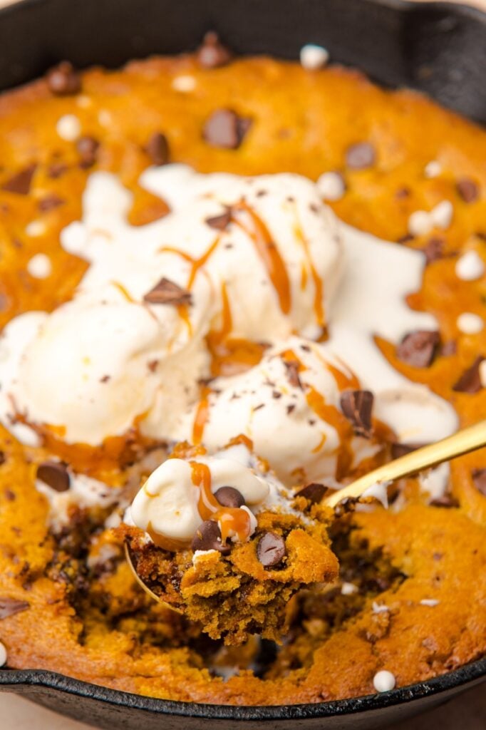 A close-up of a skillet cookie topped with melting vanilla ice cream, drizzled with caramel sauce, and sprinkled with chocolate chips and white chocolate pieces, with a spoonful being scooped out.