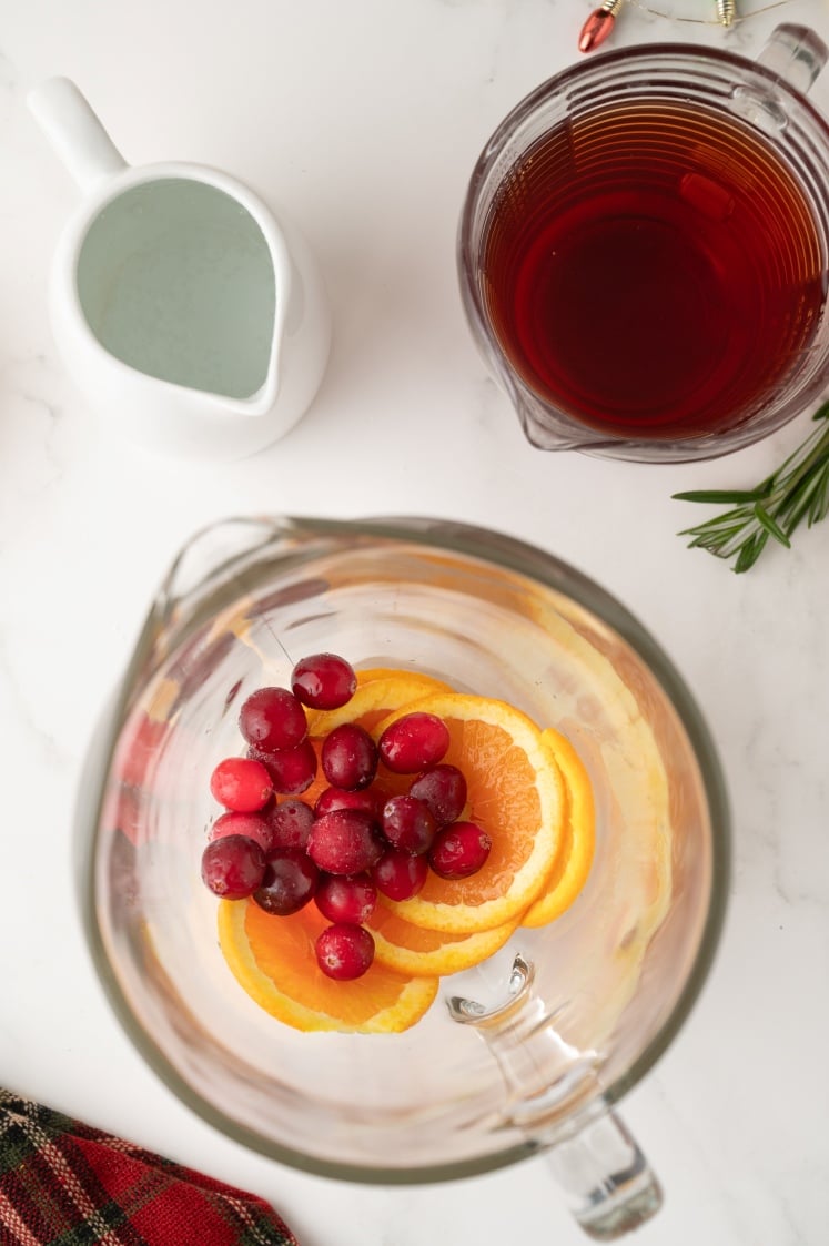 Top-down view of a pitcher with orange slices and cranberries, next to a small white jug of liquid and a measuring pitcher filled with red juice.