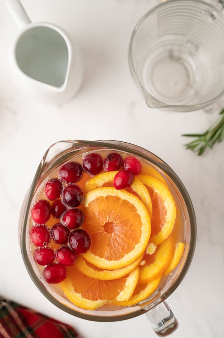 Top-down view of a pitcher filled with Christmas punch, garnished with fresh cranberries and orange slices, with an empty measuring pitcher and a white jug in the background.