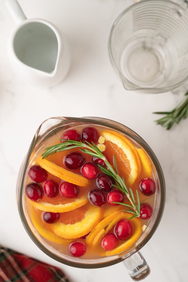 Top-down view of a pitcher of Christmas punch filled with orange slices, cranberries, and a sprig of rosemary, with an empty measuring pitcher and a white jug in the background.