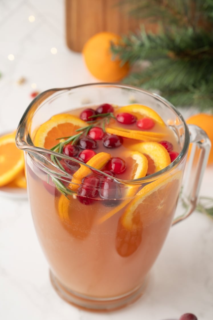 A clear glass pitcher filled with festive holiday punch, garnished with orange slices, fresh cranberries, and sprigs of rosemary, set on a light countertop with oranges and evergreen branches in the background.