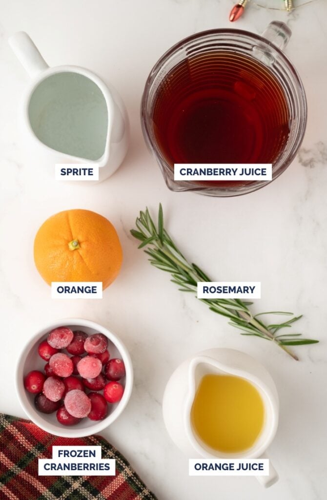 Top-down view of Christmas punch ingredients arranged on a white surface, including Sprite, cranberry juice, an orange, fresh rosemary, frozen cranberries, and orange juice, each labeled with text.