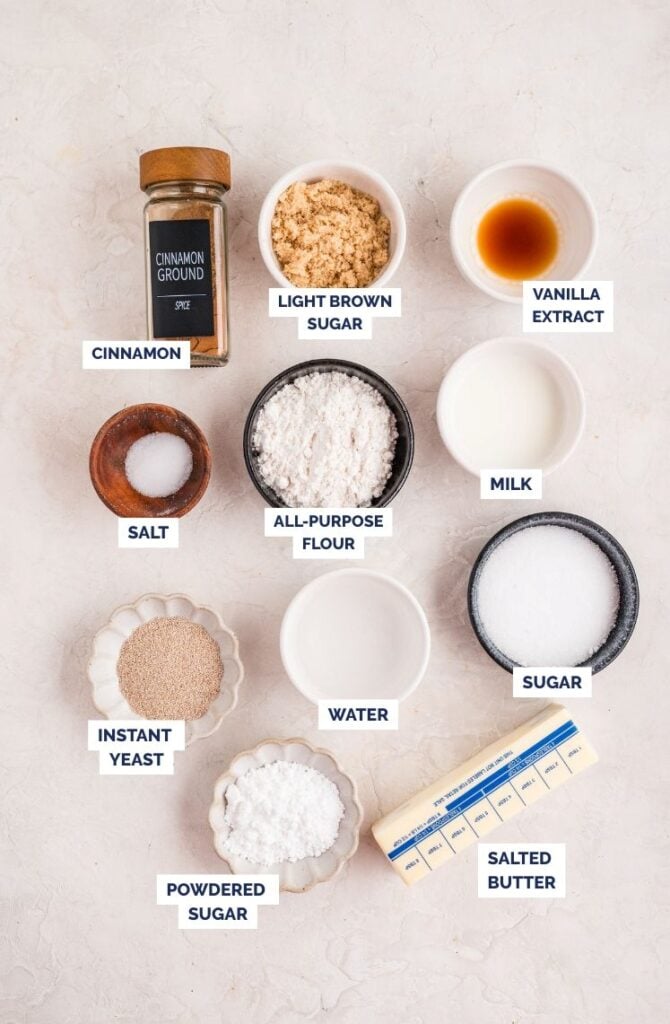 Flat lay of labeled baking ingredients arranged on a light surface, including a jar of ground cinnamon, bowls of light brown sugar, vanilla extract, milk, all-purpose flour, salt, instant yeast, sugar, powdered sugar, water, and a stick of salted butter.