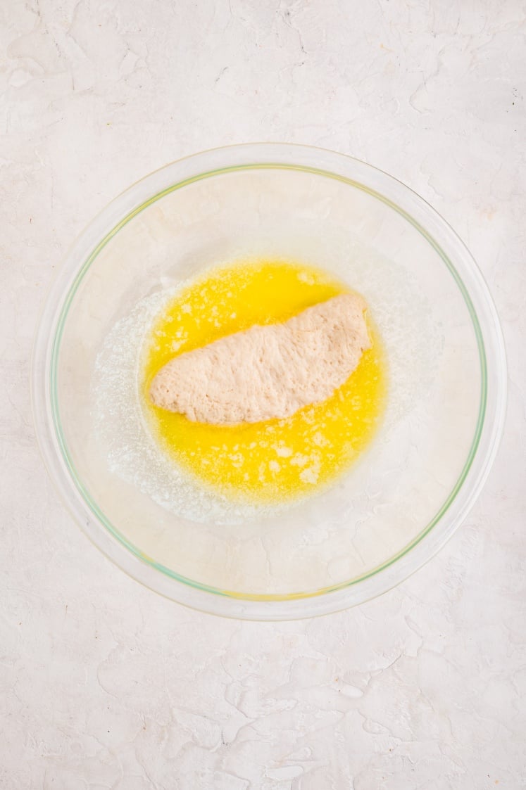 A single piece of dough placed in a glass bowl filled with melted butter, preparing it to be coated before adding the cinnamon-sugar mixture.
