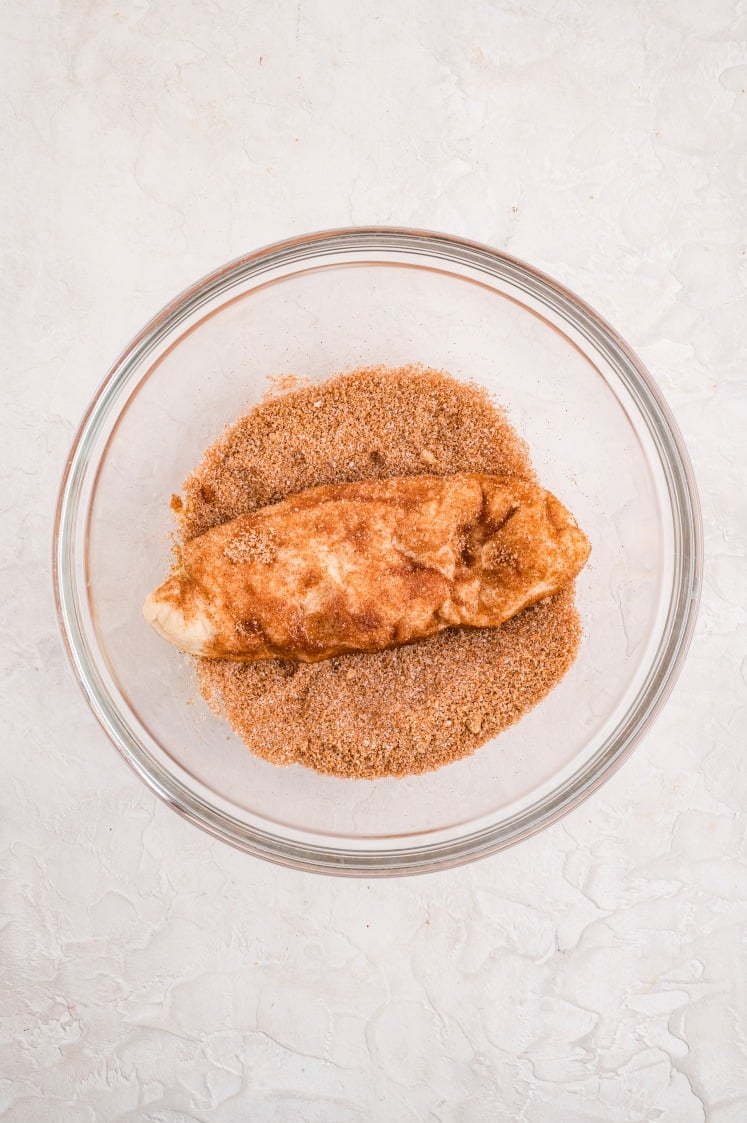 A piece of dough coated in cinnamon-sugar mixture inside a glass bowl, showing the process of covering each dough section for pull-apart bread.