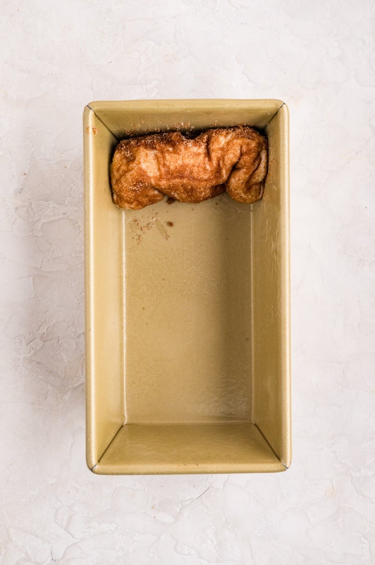 A small piece of dough covered in cinnamon sugar, ready to become delicious cinnamon pull apart bread, sits in the corner of an empty rectangular metal loaf pan.
