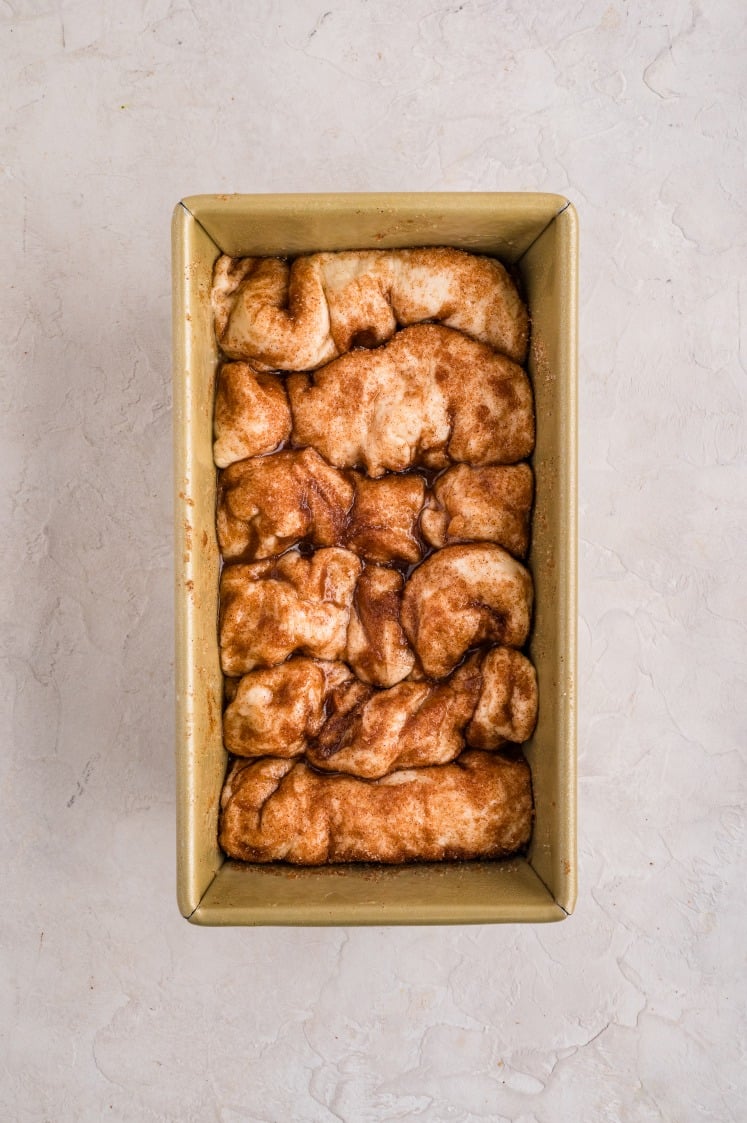 A rectangular loaf pan filled with unbaked cinnamon pull apart bread dough sits on a light-colored surface, ready to become a delicious treat.