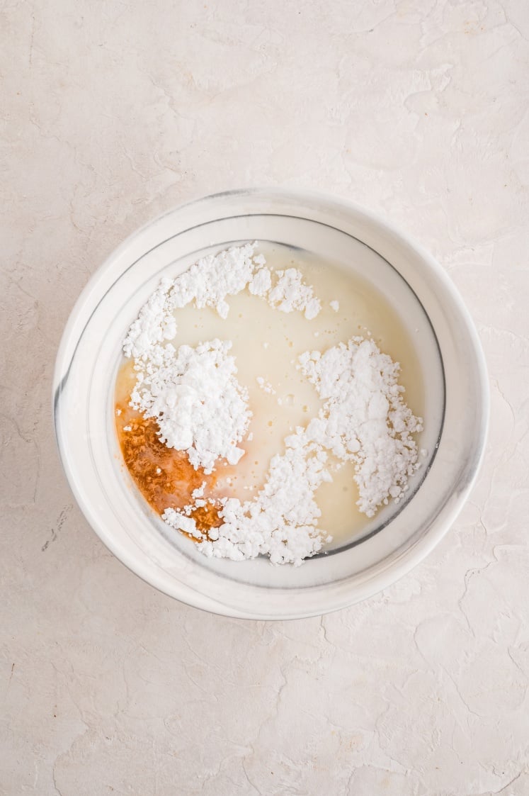 A white bowl containing powdered sugar, milk, and vanilla extract on a light textured surface—perfect for glazing cinnamon pull apart bread.