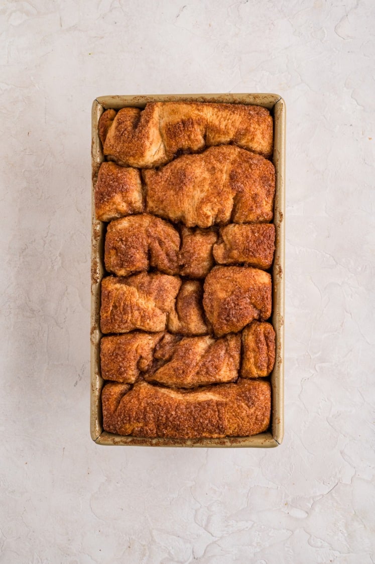 A loaf pan filled with golden-brown cinnamon pull apart bread pieces coated in cinnamon sugar, shown from above on a light textured surface.