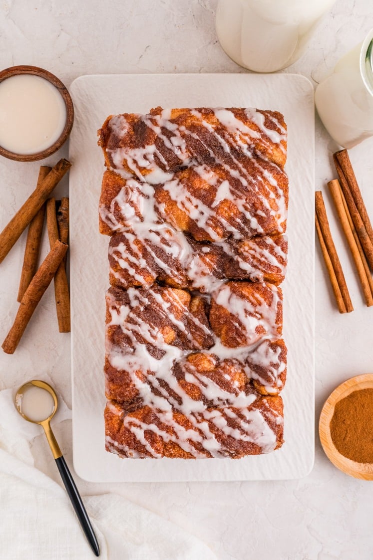 A rectangular loaf of cinnamon pull apart bread with white icing drizzle sits on a white platter, surrounded by cinnamon sticks, a jar of milk, and a small bowl of cinnamon.