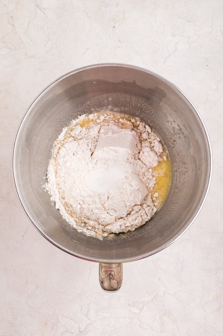 A stainless steel mixing bowl filled with flour, sugar, and other dry ingredients sitting on top of a melted butter mixture, ready to be combined into dough.