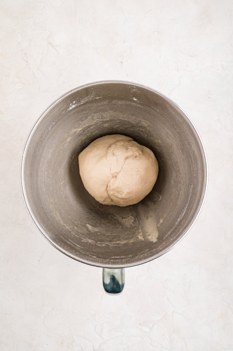 A smooth ball of dough resting in the center of a stainless steel mixing bowl after being fully kneaded and formed.