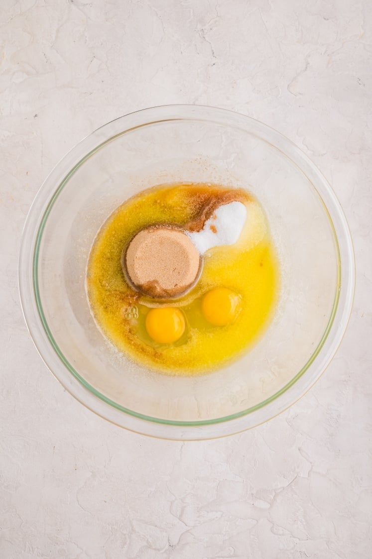 A glass mixing bowl filled with melted butter, two cracked eggs, brown sugar, granulated sugar, and vanilla extract, ready to be whisked together for birthday cake brownies.