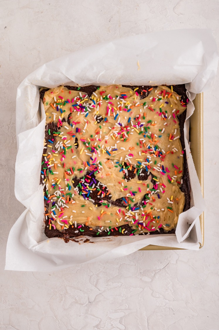 Unbaked birthday brownies in a parchment-lined pan, topped with swirls of funfetti cake batter and sprinkled with colorful rainbow sprinkles before baking.