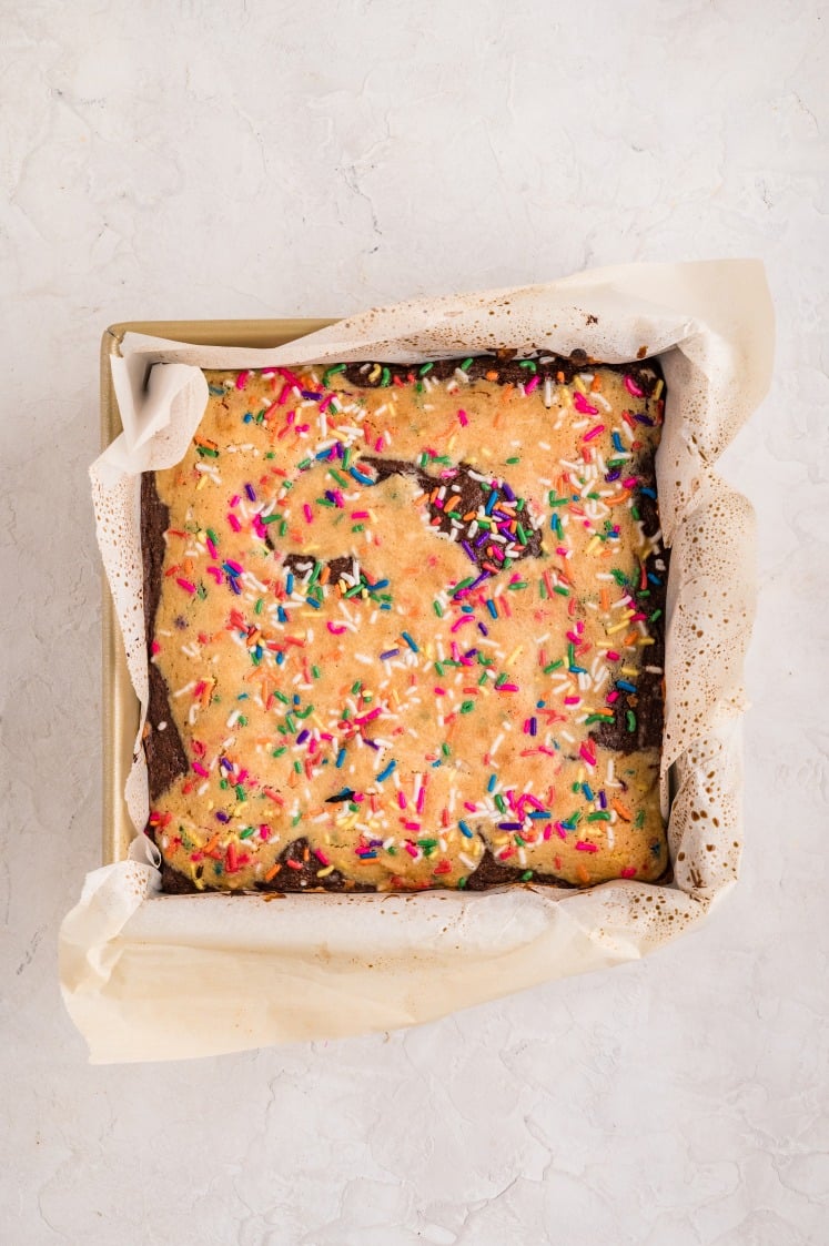 A pan of freshly baked birthday cake brownies lined with parchment paper, showing a golden funfetti cookie layer swirled with chocolate brownie and topped generously with rainbow sprinkles.